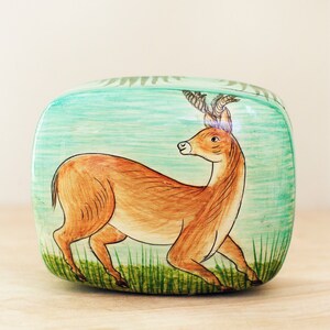 May include: A rectangular wooden box with a painted design of a deer with antlers standing in a grassy field. The background is a light blue color.