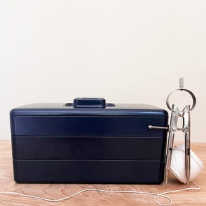 May include: A navy blue, three-tiered toolbox with a handle, next to a silver compass and a ball of white string. The toolbox is on a wooden surface.