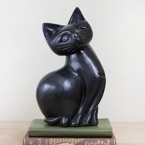 May include: A glossy black cat sculpture with a seated pose, perched atop two vintage books. The cat has a sleek, rounded body and pointed ears. The books are bound in green and brown with gold lettering.