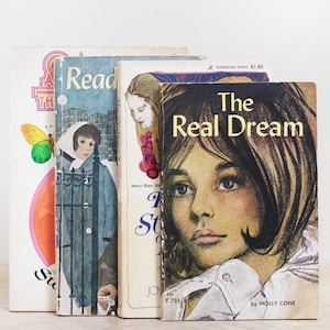 May include: A stack of four vintage paperback books. The books have colorful covers with illustrations and text. Titles include "The Real Dream" and "Read". The books are stacked vertically, with the spines facing forward.