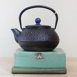 May include: A black cast iron teapot with a textured surface and a curved handle. The teapot has a blue knob on the lid and sits atop a stack of a green wooden box and a green book.