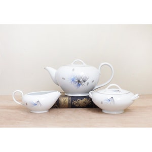 May include: A white teapot, creamer, and sugar bowl with a blue and silver pine cone design. The set is sitting on a vintage book.