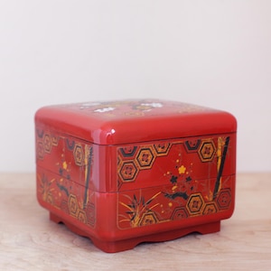 May include: A red, square, lacquered Japanese bento box with a glossy finish. The box features intricate designs of geometric patterns, bamboo, and floral motifs in black, gold, and white. The box is sitting on a light-colored wooden surface.