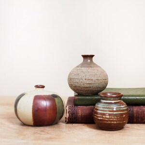 May include: Three small ceramic vases of varying shapes and finishes. One vase features wide vertical stripes in cream, brown, and green. Another is a textured, light-colored sphere. The third is a small, brown, horizontally ribbed vessel.