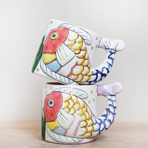 May include: Two ceramic mugs stacked, each featuring a colorful fish design. The fish have red, yellow, and blue details, with a tail-shaped handle. The mugs are white with green accents and sit on a light wooden surface.