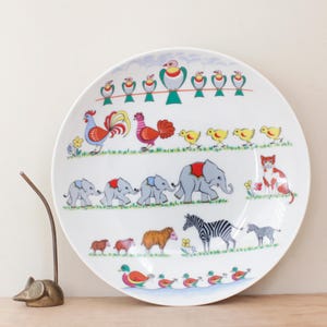 May include: White ceramic plate with a colorful illustration of various animals, including birds, chickens, elephants, a cat, zebras, and ducks. The plate has a whimsical design with a light blue border and is suitable for serving food.