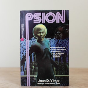 May include: A vintage paperback book titled "PSION" by Joan D. Vinge. The cover features a futuristic illustration of a person in a dark blue outfit with a yellow-blonde hairstyle, set against a dark background with the text "Cat's caught up in a chilling government experiment."