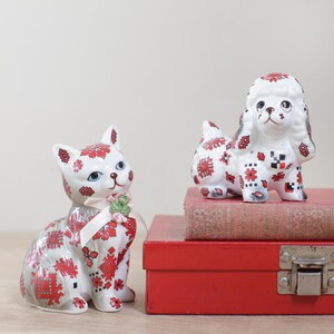 May include: Two ceramic figurines, a cat and a poodle, both decorated with a white and red geometric pattern. The poodle is sitting on a stack of books, and the cat is sitting on a wooden surface.