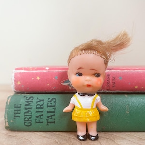 May include: A vintage doll with blonde hair, wearing a white top with yellow straps and a yellow skirt, black shoes. The doll is standing in front of two books, one green with "The Grimm's Fairy Tales" and one pink with stars.