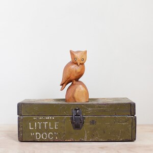 May include: A wooden owl figurine perched on a wooden base, sitting on top of a green metal box with the text "SCHAF/HENS LITTLE "DOC"' printed on the front.