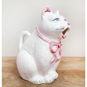 May include: White ceramic cat-shaped pitcher with blue eyes and a pink bow. The pitcher has a handle and a spout shaped like an open mouth. The cat is wearing a pink ribbon around its neck.