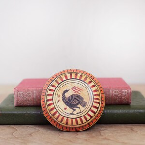 May include: A round, decorative box with a turkey illustration in the center. The box has a cream-colored background with a black turkey design. The rim is orange with red accents. The box is placed on top of two books.