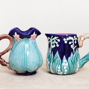 May include: Two ceramic pitchers with floral designs. One pitcher is blue with white flowers and green leaves. The other pitcher is blue with pink flowers and green leaves.