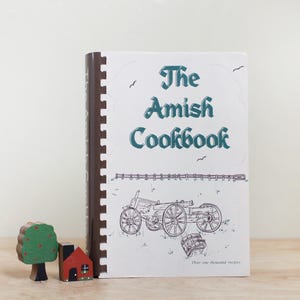 May include: A cookbook titled "The Amish Cookbook" with a white cover and teal lettering. The cover features a line drawing of a horse-drawn cart and a barrel. The binding is brown with a spiral design. A small wooden tree and house are in the foreground.