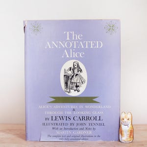 May include: A hardcover book titled The Annotated Alice by Lewis Carroll with a light purple cover featuring a central black and white illustration of Alice, green decorative accents, and text detailing the contents and contributors.