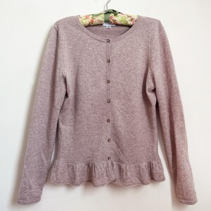 May include: A light mauve cardigan with a round neckline and long sleeves. The button-down sweater has a ruffled hem and is hanging on a floral print hanger. The buttons are a light brown color.