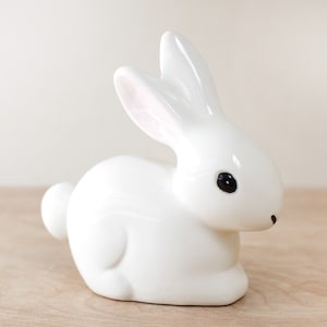 May include: A white ceramic bunny figurine with black eyes. The bunny is sitting on a wooden surface.