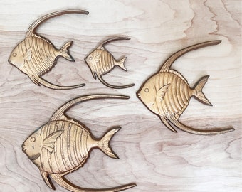 May include: Four gold-colored metal fish wall decor pieces in different sizes. The fish are stylized with a curved fin and a striped pattern. The fish are mounted on a light wood background.