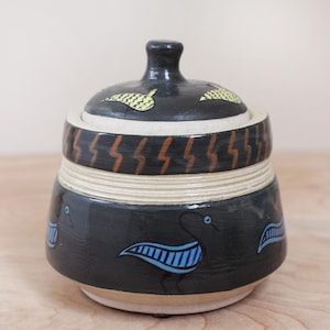 May include: A ceramic jar with a lid, decorated with stylized birds and geometric patterns in blue, yellow, brown, and white.