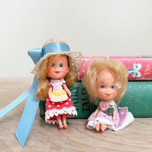 May include: Two small vintage dolls. One doll wears a straw hat with a blue ribbon and a red polka dot dress. The other doll is dressed in a pink dress and sits on a white pillow. Both dolls have blonde hair and rosy cheeks.