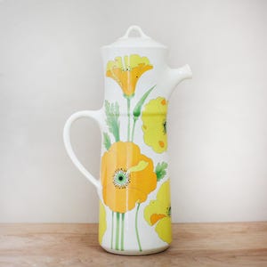 May include: White ceramic pitcher with a lid, handle, and spout. The pitcher is decorated with large yellow and orange poppy flowers and green leaves. The flowers have detailed centers. The pitcher is on a wooden surface.
