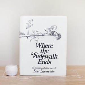 May include: A white book with a black and white illustration of a girl and a dog on the cover. The title of the book is "Where the Sidewalk Ends" and the author is Shel Silverstein.