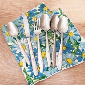 May include: A set of vintage silverware, including spoons, a fork, and a knife, arranged on a blue and white floral patterned napkin. The silverware has a starburst design on the handles. The napkin is on a wooden surface.