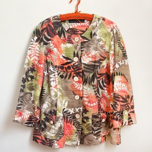 May include: A colorful button-down jacket with a botanical print. The jacket features a mix of orange, green, brown, and white leaf and floral patterns. It has a V-neck, three-quarter sleeves, and large, round, light-colored buttons. The jacket is hanging on an orange hanger.
