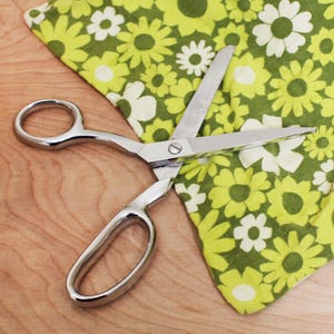 May include: Shiny, silver scissors cutting a green and white floral patterned fabric. The scissors are open, with the blades positioned to cut through the fabric. The fabric is a bright green with white and yellow daisy-like flowers.