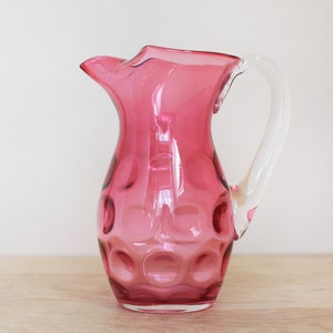 May include: A pink glass pitcher with a clear handle and a textured, bubbled design. The pitcher is perfect for serving beverages.