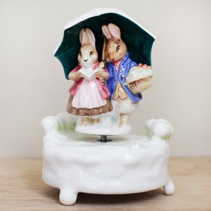May include: A porcelain musical figurine of two rabbits under an umbrella. The rabbits are dressed in colorful clothing and are holding a basket of flowers. The base of the figurine is white and has a swirling design.