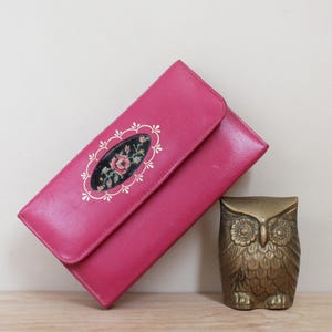 May include: A pink leather wallet with a floral embroidered design. The wallet is closed and has a flap.