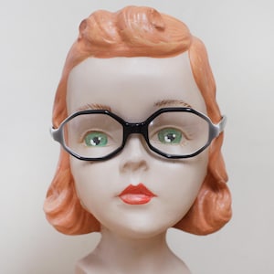 May include: A vintage-style mannequin head with red hair, green eyes, and red lips, wearing black-framed eyeglasses. The mannequin is painted with a peach skin tone and is set against a plain background.