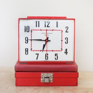 May include: A rectangular General Electric Telechron alarm clock with a red plastic frame and a white face featuring black numerals. The clock has a red and white dashed border, black hands, and a red second hand. It sits atop a red metal box.