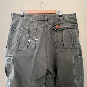 Może przedstawiać: Spodnie cargo w ciemnozielonym kolorze z dużą kieszenią po prawej stronie i mniejszą kieszenią po lewej stronie. Mniejsza kieszeń ma niewielkie rozdarcie w materiale.