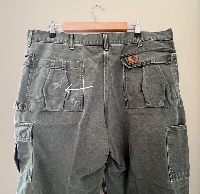 Może przedstawiać: Spodnie cargo w ciemnozielonym kolorze z dużą kieszenią po prawej stronie i mniejszą kieszenią po lewej stronie. Mniejsza kieszeń ma niewielkie rozdarcie w materiale.