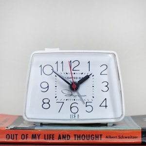 May include: A white rectangular analog alarm clock with a clear plastic frame. The clock face features black Arabic numerals, black hour and minute hands, a red second hand, and the text "Ingraham" and "LIGHTED DIAL" printed on the center.