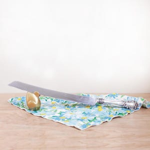 May include: A silver cake knife with a decorative handle rests on a floral patterned cloth. A small gold-colored mouse-shaped object is positioned near the knife. The cloth features a blue and yellow daisy design.