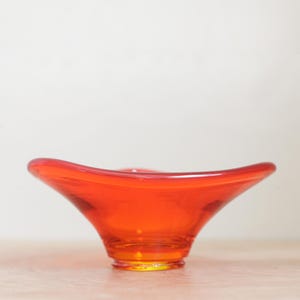 May include: A vibrant orange glass bowl featuring a wide, flared rim and a smooth, translucent finish. The bowl has a circular base that tapers upward into a shallow, open basin with gently undulating edges, creating a fluid and symmetrical form.