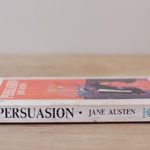 Op de afbeelding: Een paperback van Jane Austens "Persuasion" wordt getoond, met de titel en de naam van de auteur prominent weergegeven. Het boek heeft een rood en zwart omslagontwerp en ligt op een houten oppervlak.