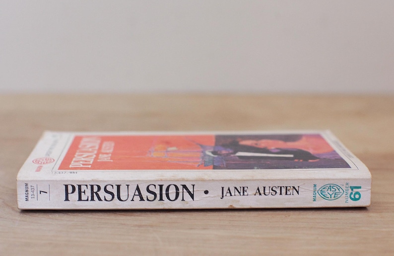 Op de afbeelding: Een paperback van Jane Austens "Persuasion" wordt getoond, met de titel en de naam van de auteur prominent weergegeven. Het boek heeft een rood en zwart omslagontwerp en ligt op een houten oppervlak.