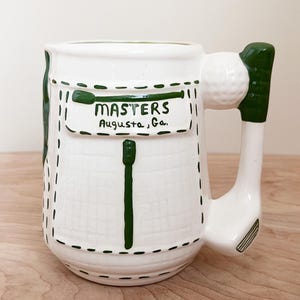 May include: White ceramic mug with a golf-themed design. The mug features a handle shaped like a golf club and a golf ball. The front of the mug has a green pocket design with the word "MASTERS" and "Augusta, Ga." printed on it.
