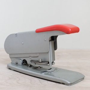 May include: A silver stapler with a red handle. The stapler is on a light wood surface.