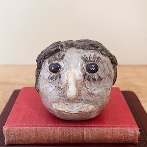 May include: A ceramic sculpture of a human head with dark hair, black eyes, and a prominent nose. The head is off-white with brown accents and sits atop a stack of red and brown books. The sculpture has a whimsical, folk art style.