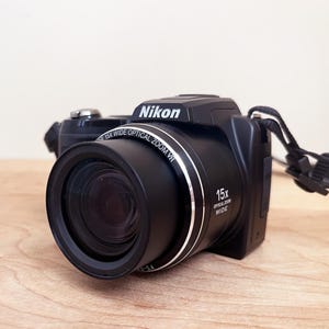 May include: Black Nikon digital camera with a 15x optical zoom lens. The camera has a built-in flash and a wrist strap. The Nikon logo is visible on the top of the camera.