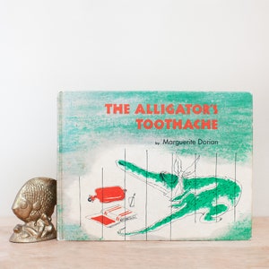 May include: A vintage children's book titled "The Alligator's Toothache" by Marguerite Dorian. The cover features a green alligator with a red hot water bottle and a glass of water. The background is a light blue and green watercolor wash.