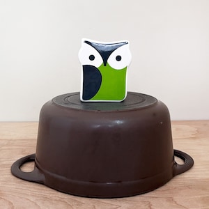 May include: A brown ceramic pot with a white owl-shaped lid. The owl has black eyes and a green body.