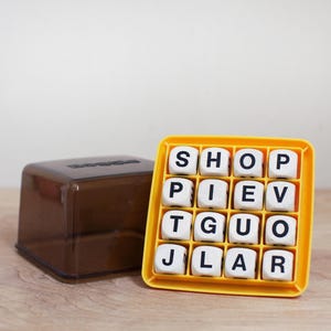 May include: A yellow word game with white letter dice and a brown plastic storage container. The dice are arranged in a grid, displaying letters like "SHOP" and "PIEV". The container is a translucent brown cube.