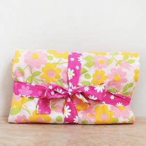 May include: A folded floral fabric item, wrapped with a bright pink ribbon tied in a bow. The fabric features a pattern of yellow, pink, and green flowers on a white background. The ribbon is adorned with white daisy-like flowers.