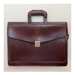 May include: A dark brown leather briefcase with a top handle and a flap closure. The briefcase has a gold-colored clasp and a rectangular shape. The handle is also brown and appears to be made of leather.